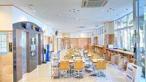 Eine großzügige Cafeteria mit langen Tischen und Stühlen, einem Verkaufsautomaten und einer Uhr an der Wand.