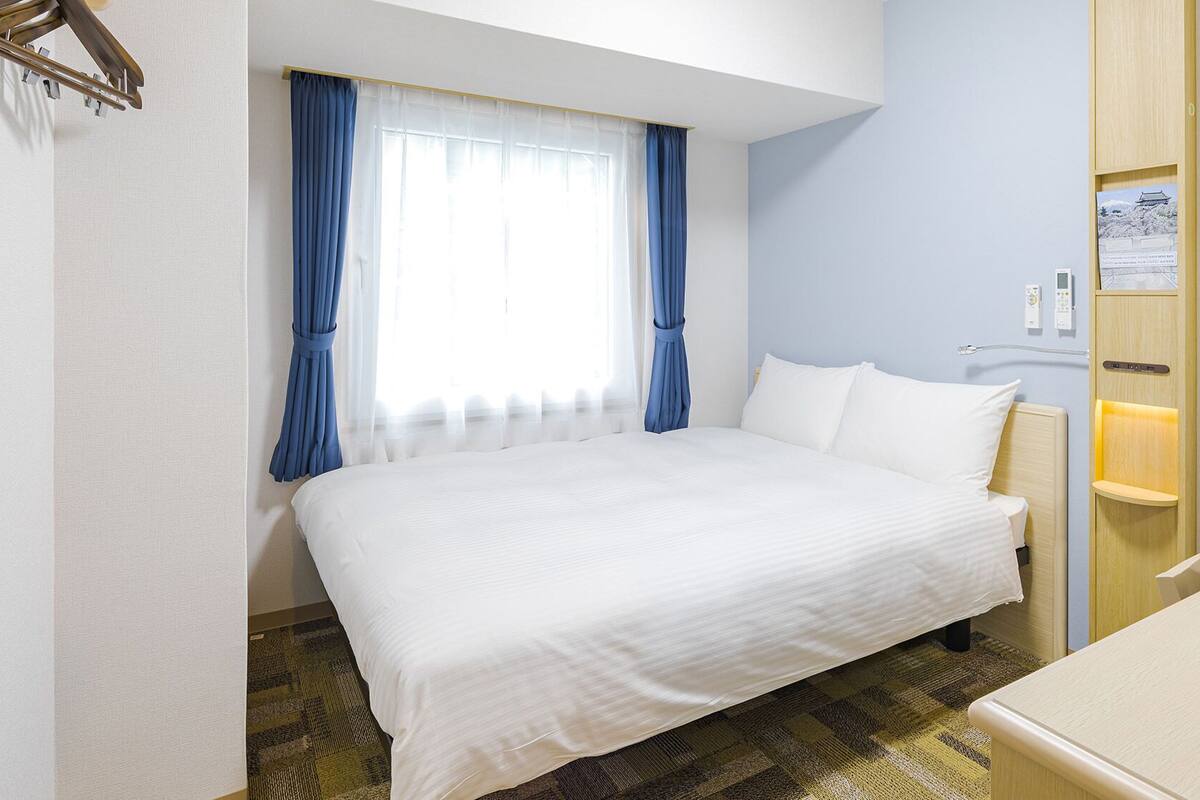Ein Hotelzimmer mit Bett, Schreibtisch, Stuhl, Nachttisch, einem Fenster mit Vorhängen und einem Wandregal mit Lampe.