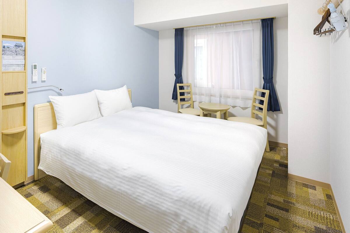Ein Hotelzimmer mit Bett, Schreibtisch, zwei Stühlen, einem kleinen Tisch und einem Vorhangfenster.