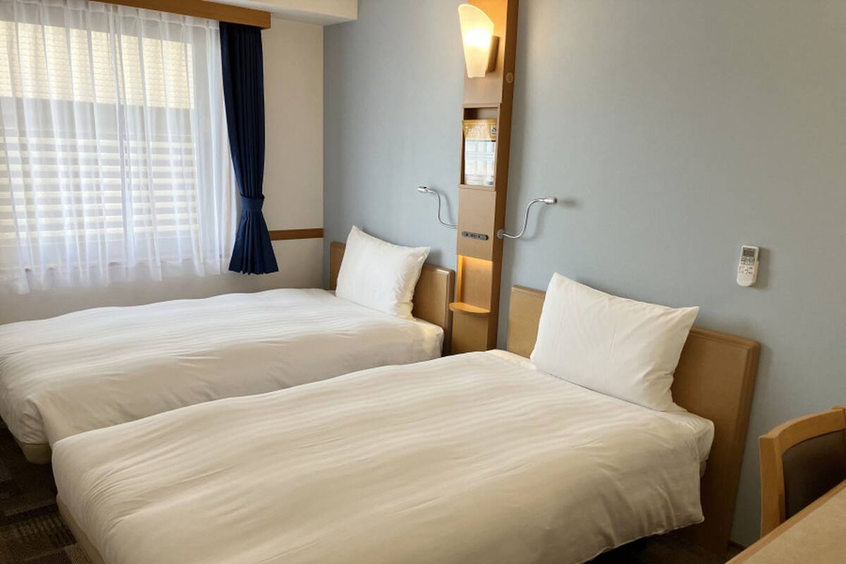 Een hotelkamer met twee bedden, een raam met dunne gordijnen, een nachtkastje en een wandlamp.