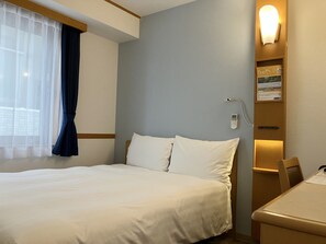 Une chambre d’hôtel comprenant un lit, un bureau, une chaise, une fenêtre avec des rideaux et un luminaire fixé au mur.