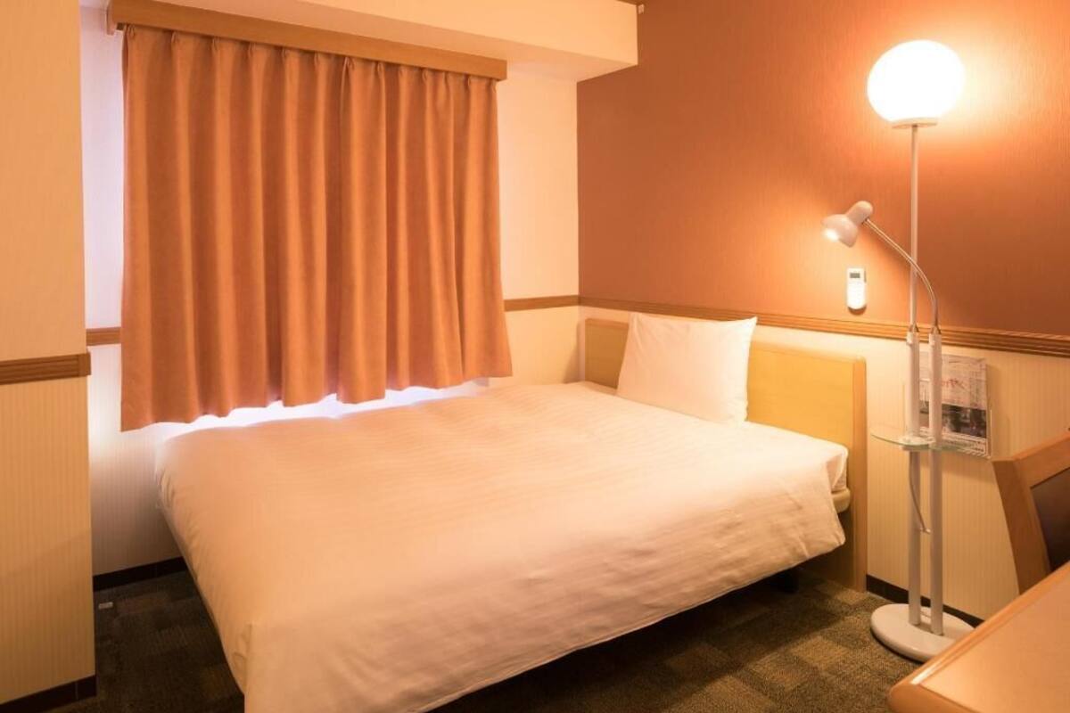 Een hotelkamer met een bed, een nachtkastje, een lamp en een raam met gordijnen.