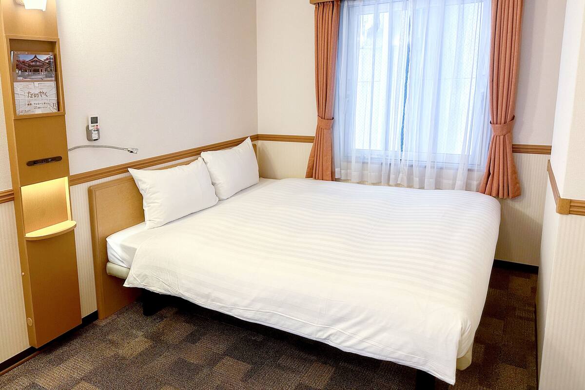 Ein Hotelzimmer mit einem großen Bett, einem Nachttisch, einem Schreibtisch, einem an der Wand befestigten Telefon und einem Fenster mit Vorhängen.