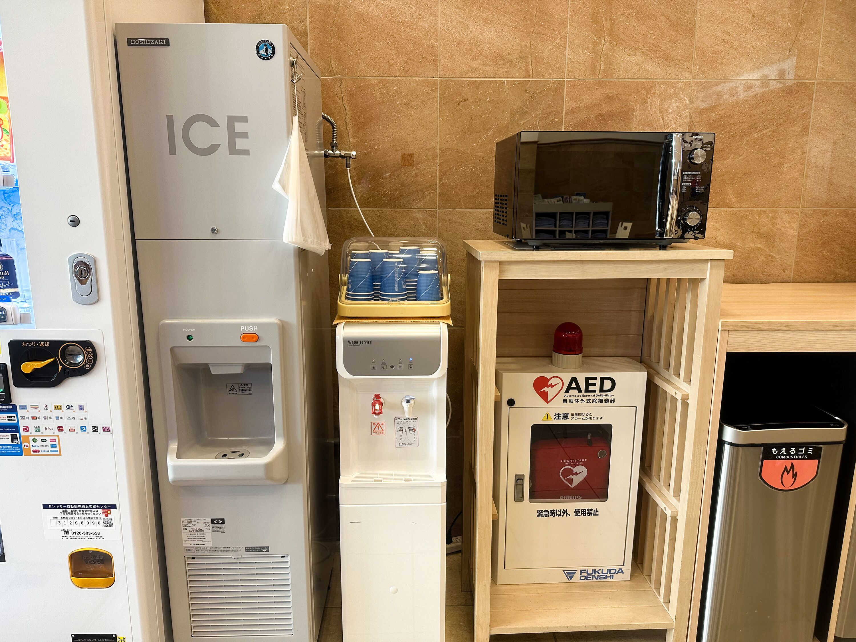 Un refrigerador con la etiqueta 'ICE', un dispensador de agua, un microondas, un botiquín de desfibrilación (DEA) y un contenedor de basura.