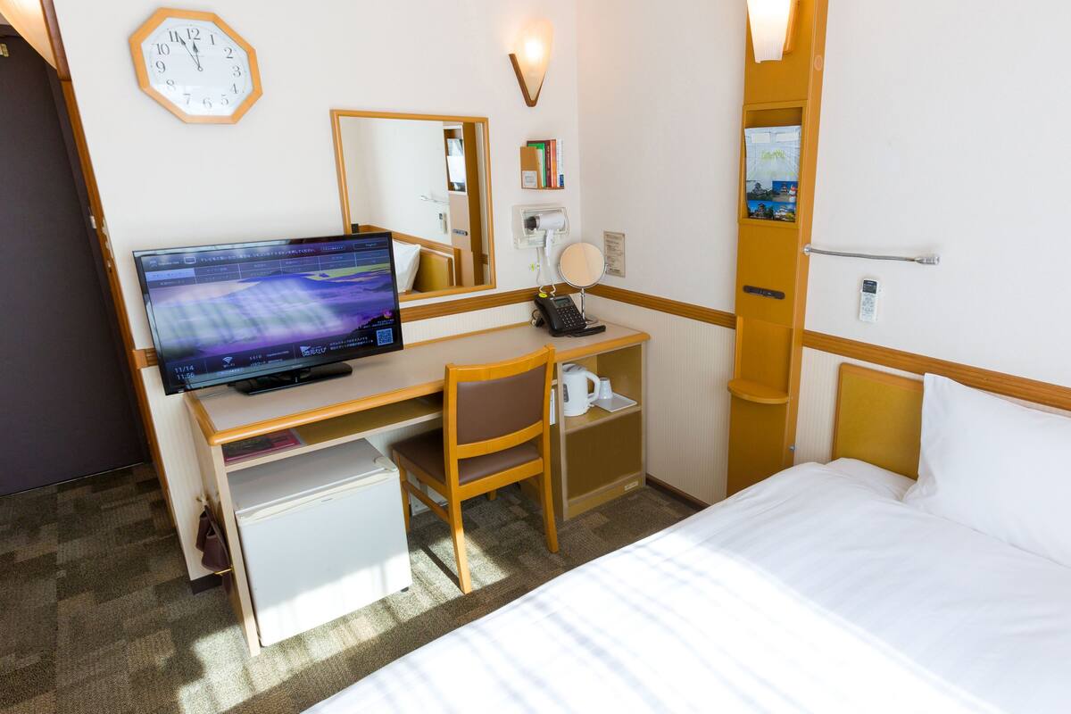 Ein Hotelzimmer mit einem Bett, einem Schreibtisch mit Computer, einem Sessel, einem kleinen Kühlschrank, einer Uhr und einem Spiegel.