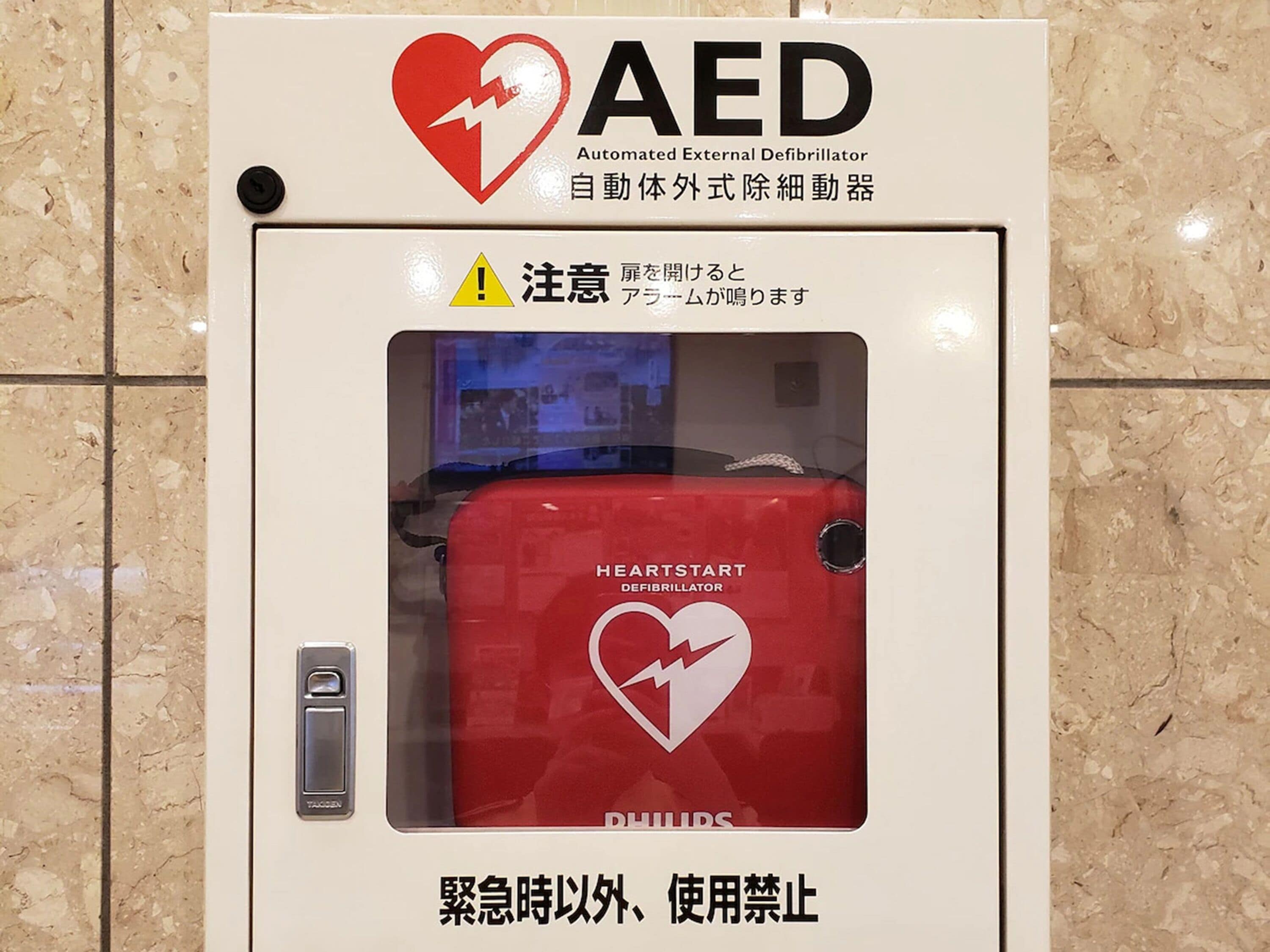 An AED (Automated External Defibrillator) box with a red heart symbol, a warning sign, and text in multiple languages.