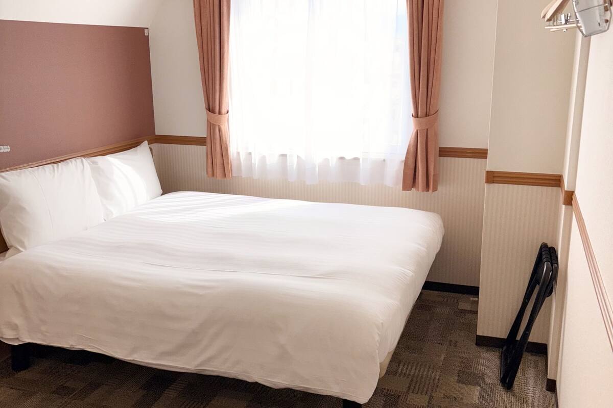 A hotel room with a large bed, a wooden headboard, a window with sheer curtains, and a wooden shoe rack.