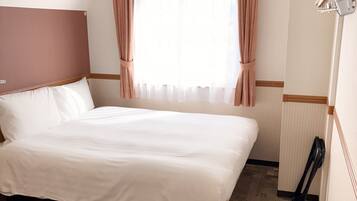 A hotel room with a large bed, a wooden headboard, a window with sheer curtains, and a wooden shoe rack.