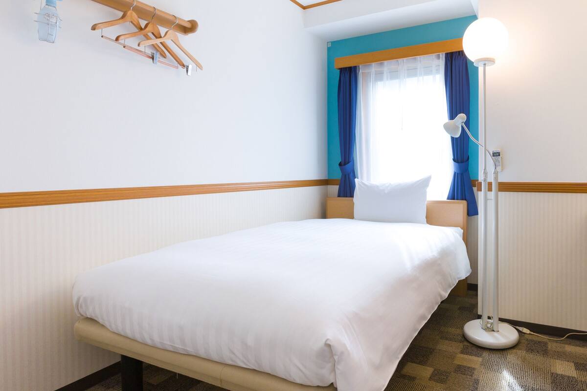 A hotel room with a bed, a bedside lamp, a window with curtains, and a wall-mounted drying rack.