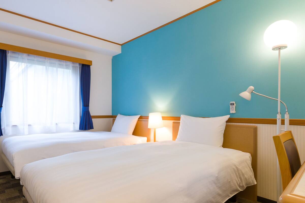 A hotel room with two beds, a desk with a lamp, and a view of the outside through a window with curtains.