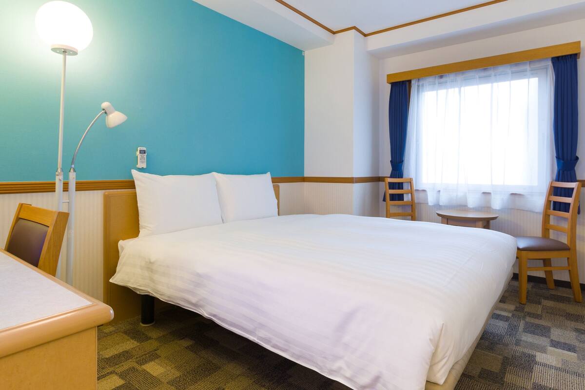 A hotel room with a large bed, a desk, a chair, a lamp, and a window with curtains.