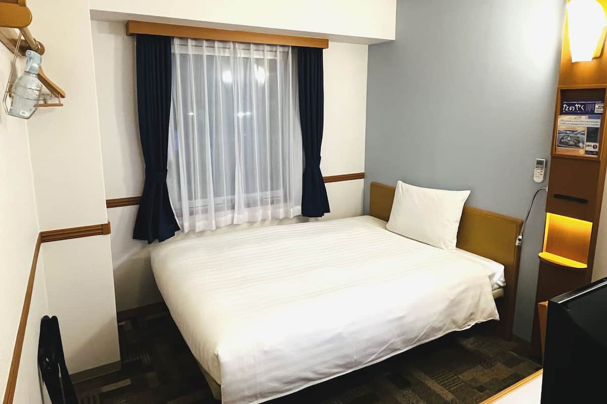 Ein Hotelzimmer mit Bett, Fenster mit durchscheinenden Vorhängen, Nachttisch mit Lampe und einem an der Wand montierten Fernseher.