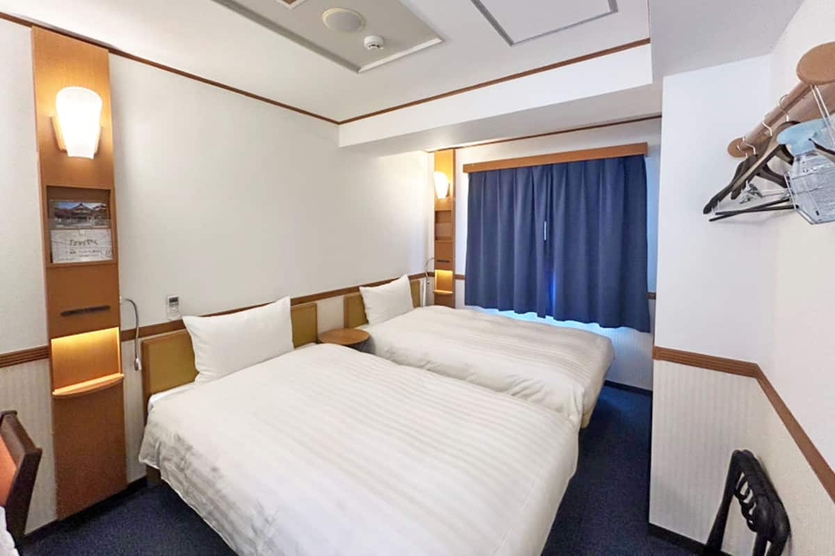 Habitación de hotel con dos camas, un escritorio, una silla y un ventanal con cortinas.