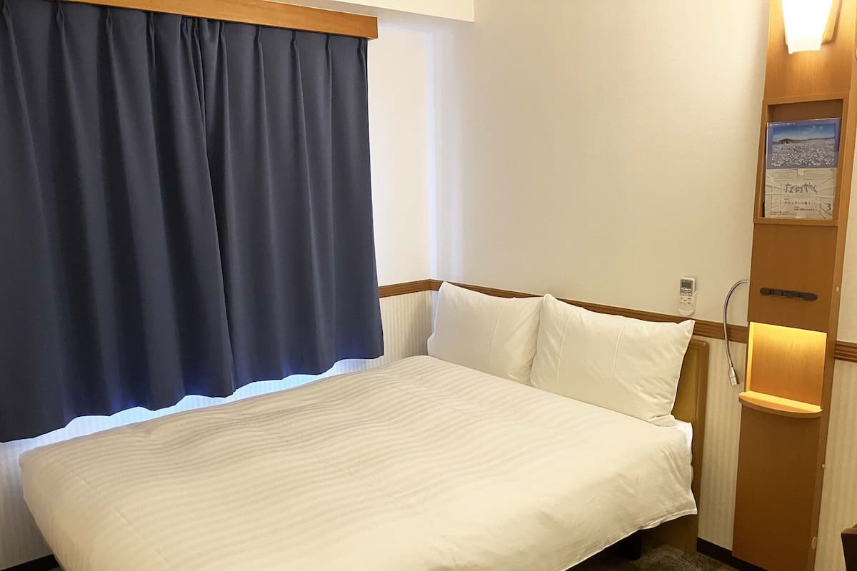 Habitación de hotel con cama, mesita de noche, lámpara y ventana con cortinas.