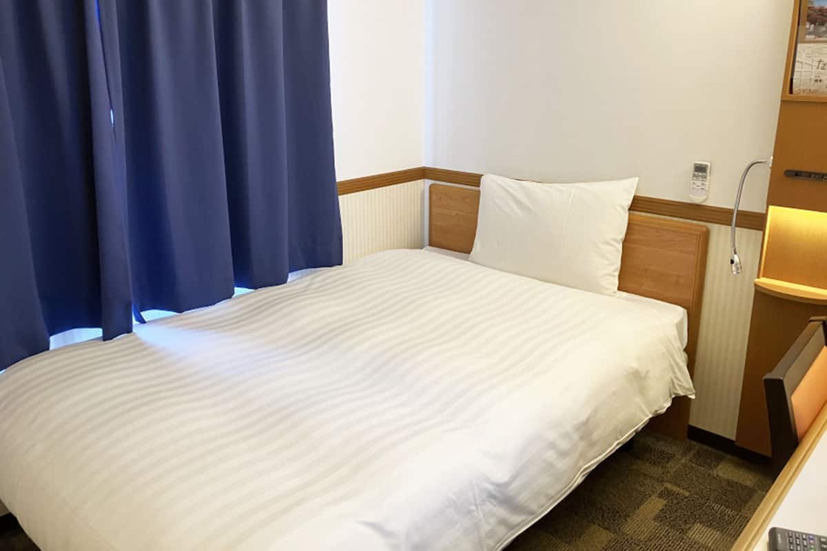 Habitación de hotel con cama, mesita de noche, lámpara, ventana con cortinas y teléfono instalado en la pared.