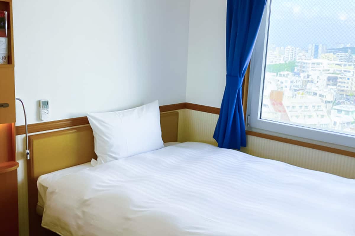 Habitación de hotel con cama, mesita de noche, teléfono y ventana con vista a un paisaje urbano.