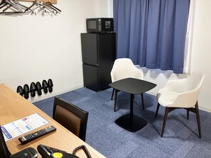 Habitación de hotel con un escritorio de madera, dos sillas blancas, una mesita negra pequeña, un refrigerador negro, un televisor encima del refrigerador, una alfombra azul y una ventana con cortinas azules.