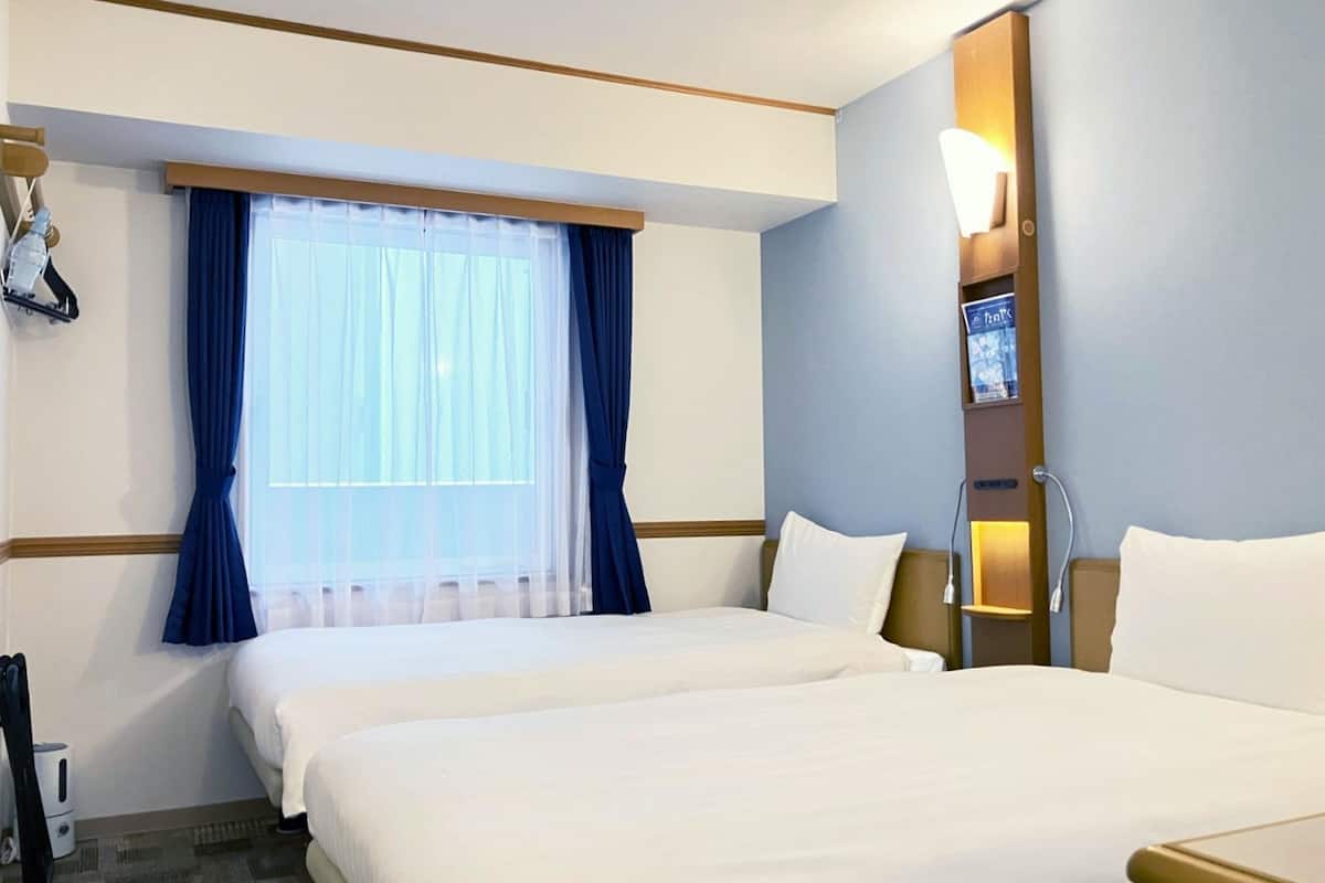 A hotel room with two beds, a large window with blue curtains, a bedside table with a lamp, and a wall-mounted TV.