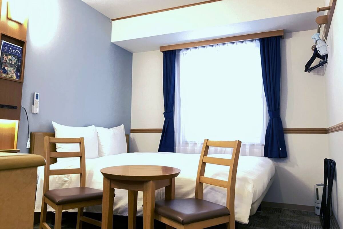 A hotel room with a bed, two chairs, a small table, a window with curtains, a coat rack, and a wall-mounted TV.