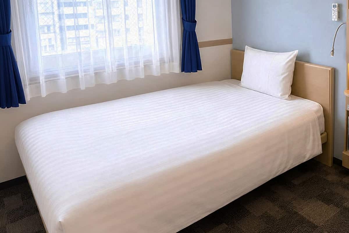 A single bed with a white mattress and pillow, a wooden headboard, a window with blue curtains, and a wall-mounted control panel.
