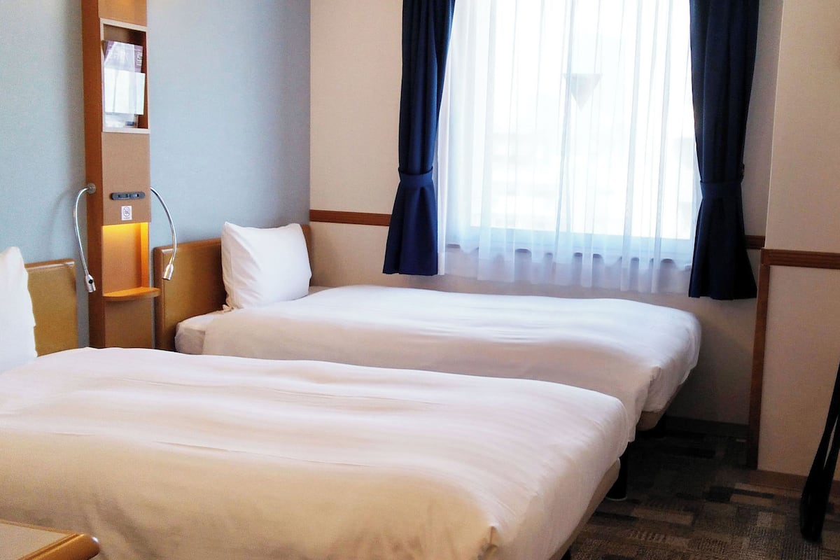 Ein Hotelzimmer mit zwei Betten, einem kleinen Schreibtisch, einem Stuhl und einem Fenster mit Vorhängen.