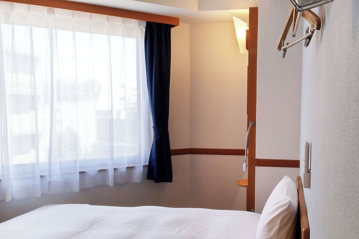 Ein Hotelzimmer mit Bett, einem Fenster mit leichtem Vorhang, einem dunklen Vorhang und einer Wandlampe.