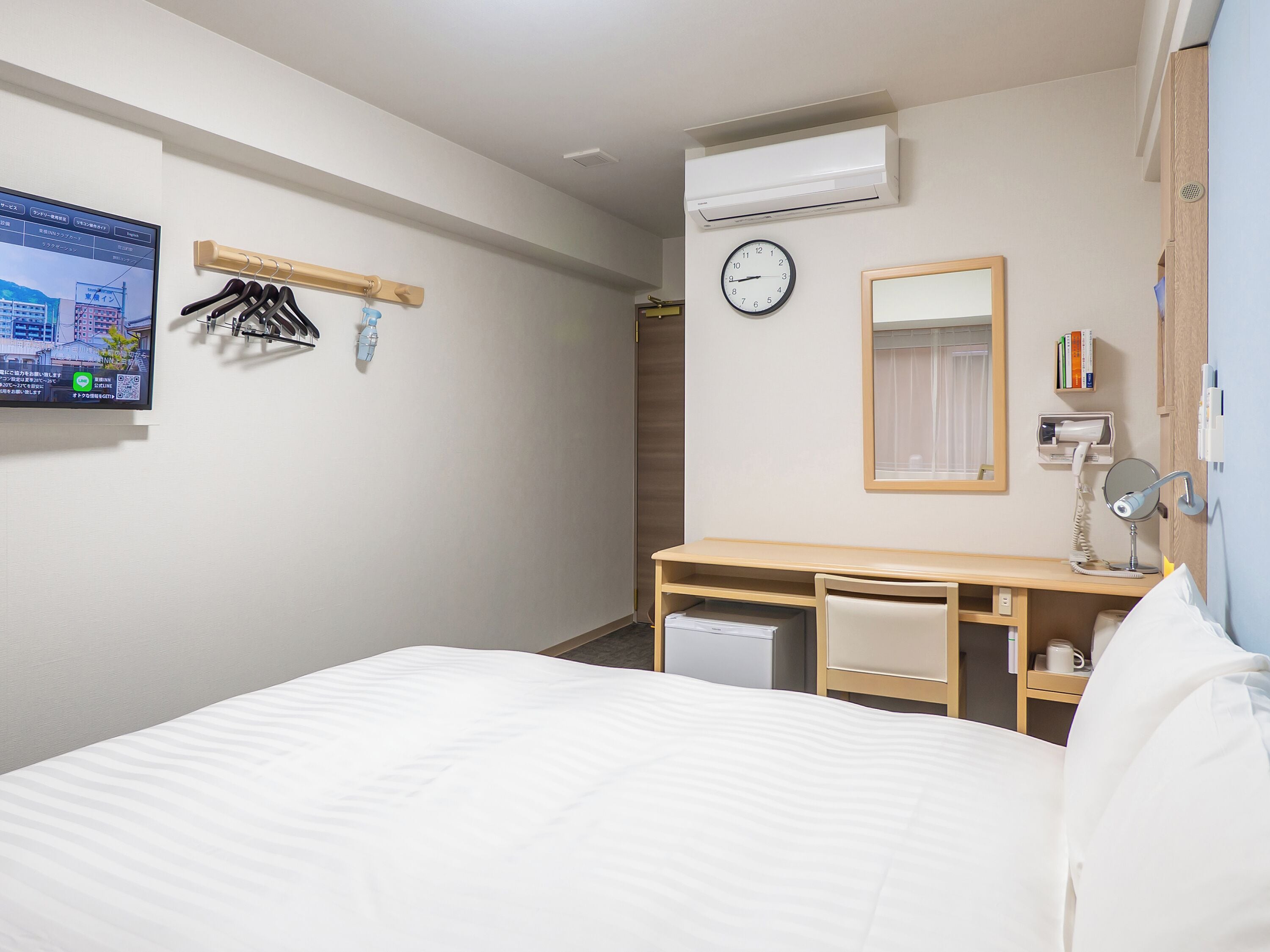 Een hotelkamer met een bed, een bureau met stoel, een kleine bureaulamp, een klok en een airconditioning.