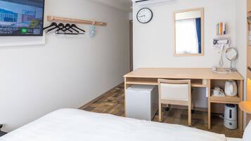 Habitación de hotel con cama, escritorio con silla, refrigerador pequeño, televisor montado en la pared, reloj, espejo, estante con artículos de aseo y perchero.