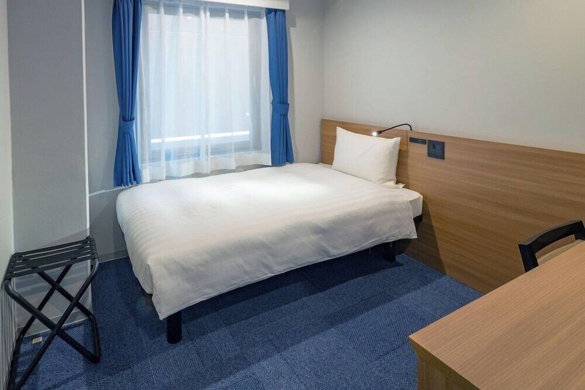 Ett hotellrum med en enkel säng, en sänggavel i trä, ett skrivbord och ett fönster med blå gardiner.