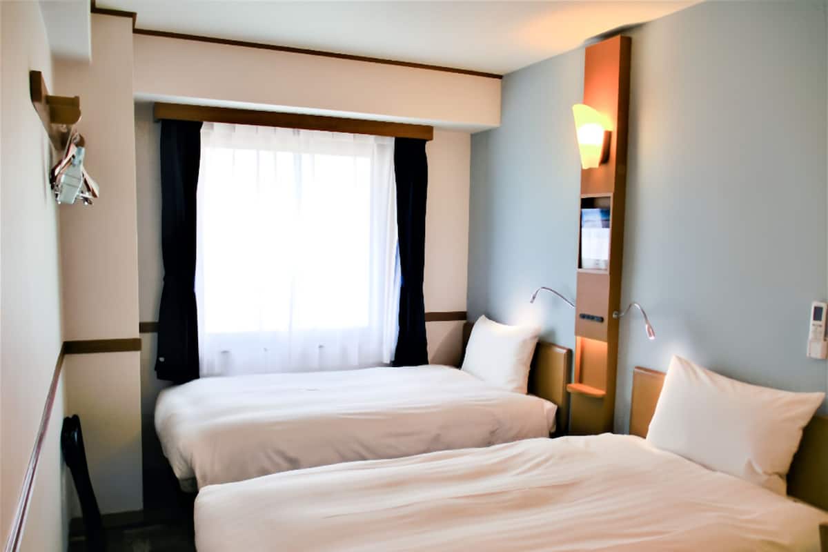A hotel room with two beds, a window with curtains, a bedside table, and a wall-mounted lamp.
