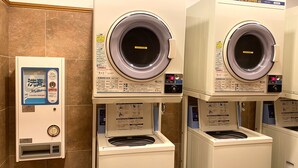 A row of identical front-loading washing machines with control panels and instruction labels, and a wall-mounted laundry detergent dispenser. - Toyoko Inn Hitachi Ekimae (Hitachi)