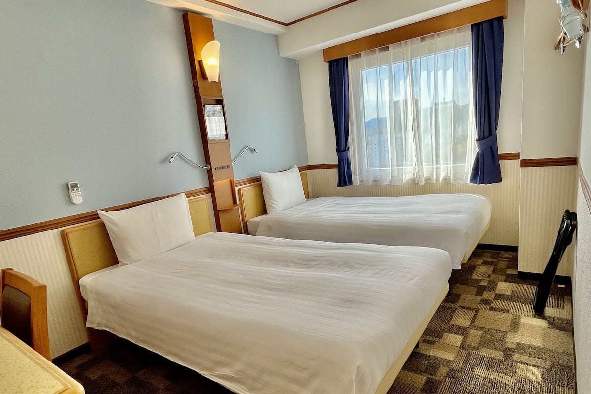 Ein Hotelzimmer mit zwei Betten, einem Fenster mit Vorhängen, einem Nachttisch und einem Spiegel.