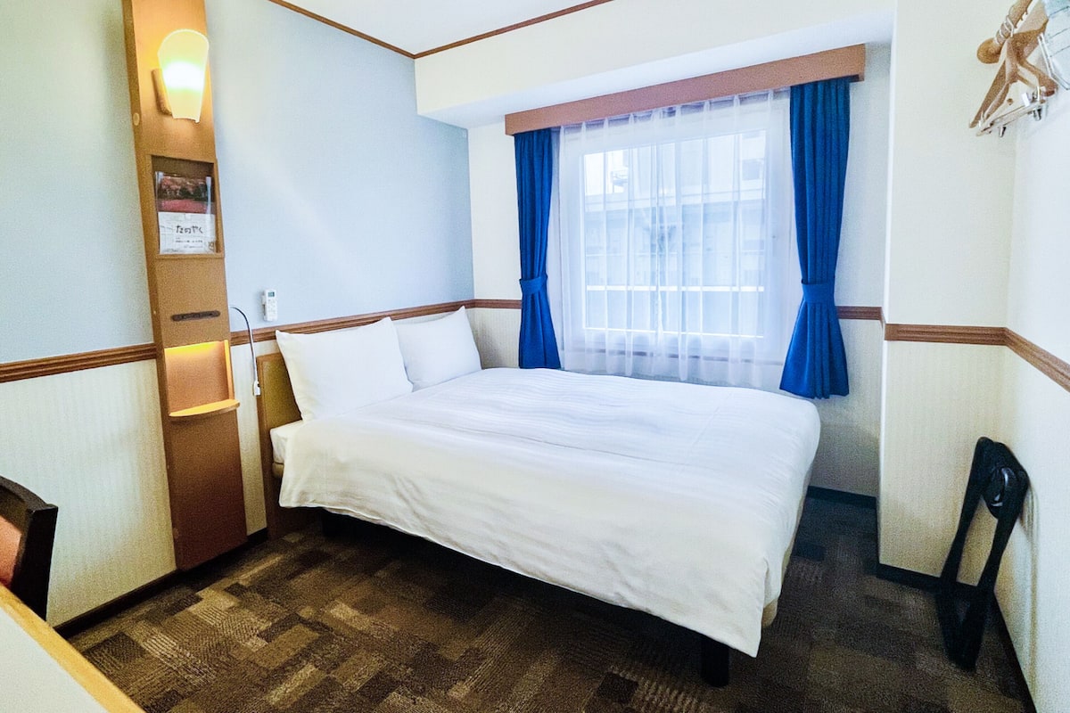 Ein Hotelzimmer mit einem Bett, einem Fenster mit blauen Vorhängen, einer Wandleuchte und einer Holzvertäfelung.