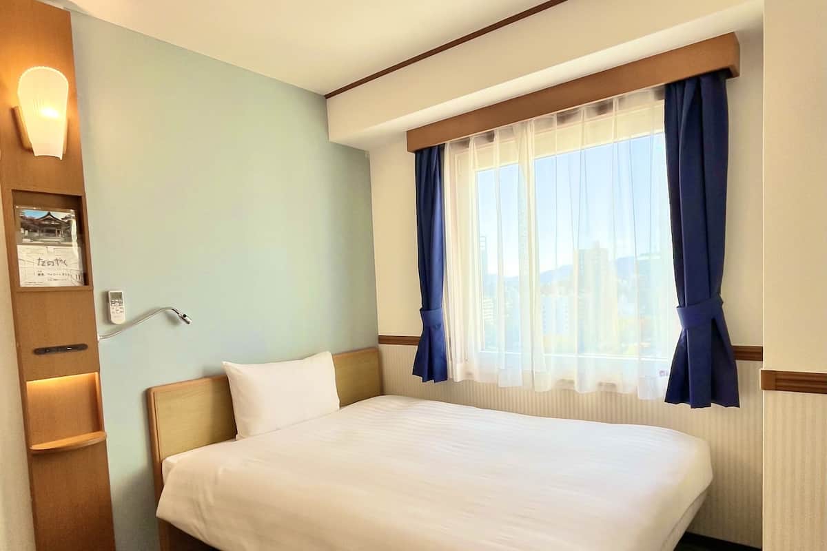 Ein Hotelzimmer mit Bett, Wandleuchte, Fenster mit Vorhängen und Bedienfeld an der Wand.