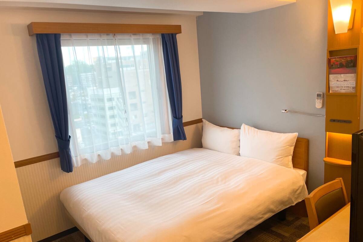 A hotel room with a bed, a window with curtains, a bedside table, and a wall-mounted TV.
