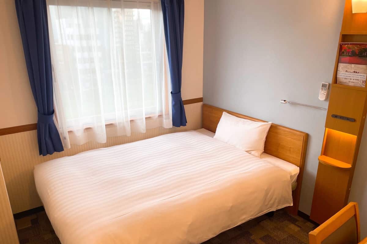 A hotel room with a large bed, a window with blue curtains, a bedside table with a lamp, and a wall-mounted shelf with a book.
