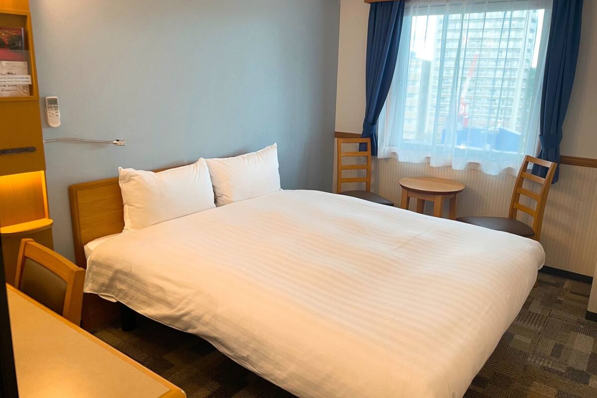 A hotel room with a large bed, a small table, two chairs, a bookshelf, and a window with curtains.