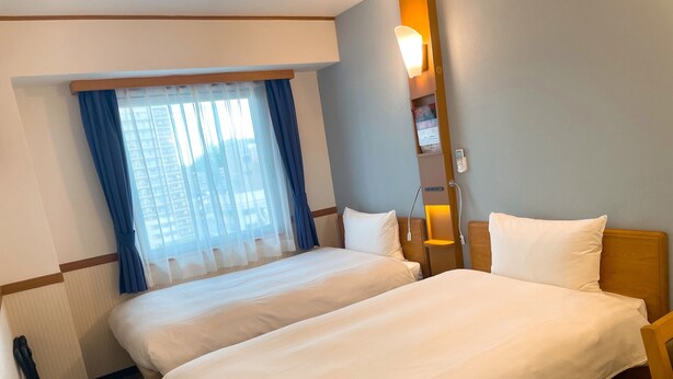 Une chambre d’hôtel avec deux lits, une fenêtre avec des rideaux bleus, une table de chevet avec une lampe et un téléphone fixé au mur.