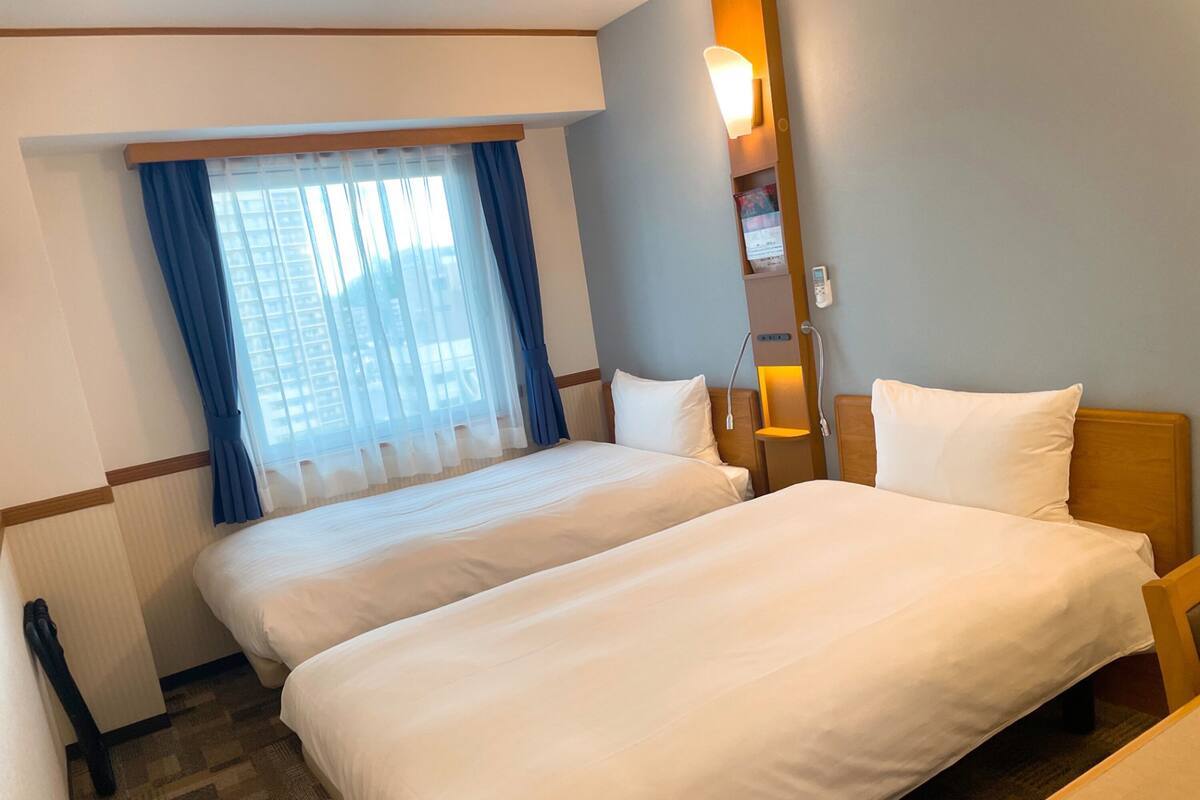 A hotel room with two beds, a window with blue curtains, a bedside table with a lamp, and a wall-mounted phone.
