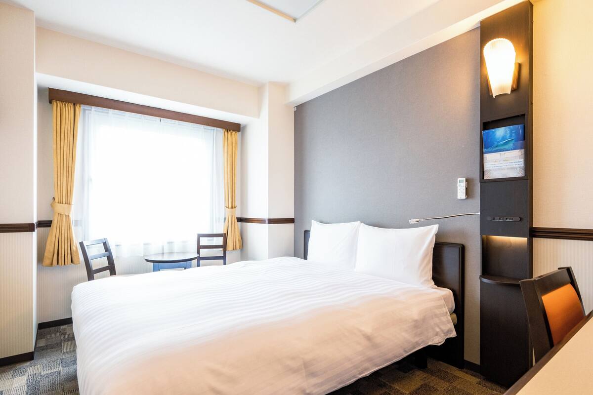 Ein Hotelzimmer mit einem großen Bett, einem kleinen Tisch, Stühlen, einem Fernseher und einer Wandleuchte.
