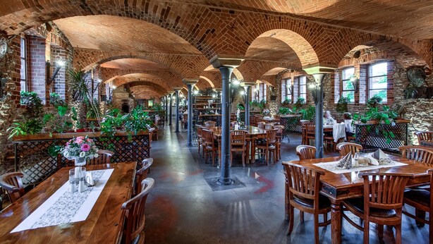 Un restaurante espacioso con arcos de ladrillo, mesas y sillas de madera, y plantas en macetas.