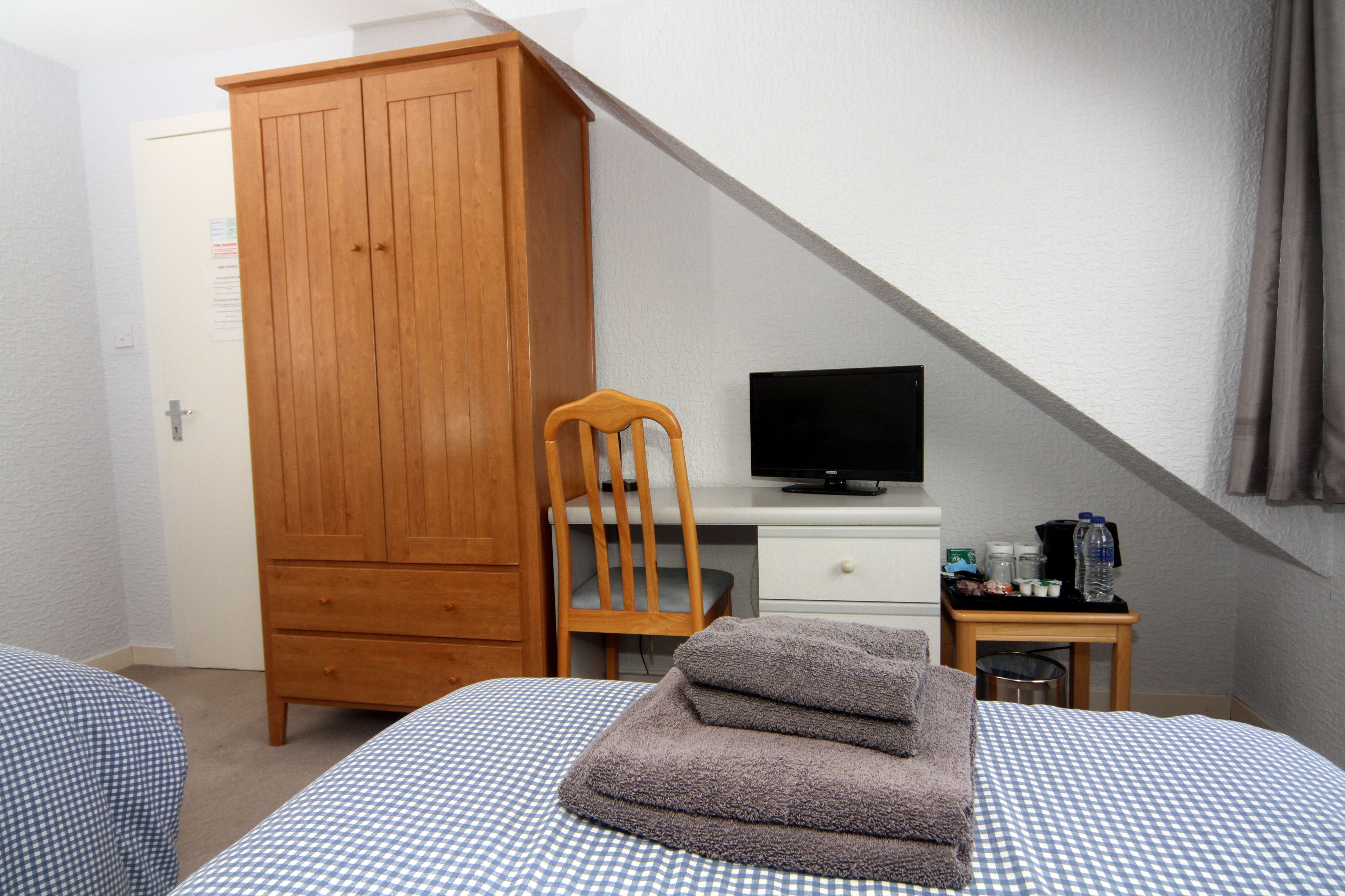 Quarto com cama, guarda-roupa de madeira, secretária com televisão e pequena mesa com artigos de higiene pessoal.