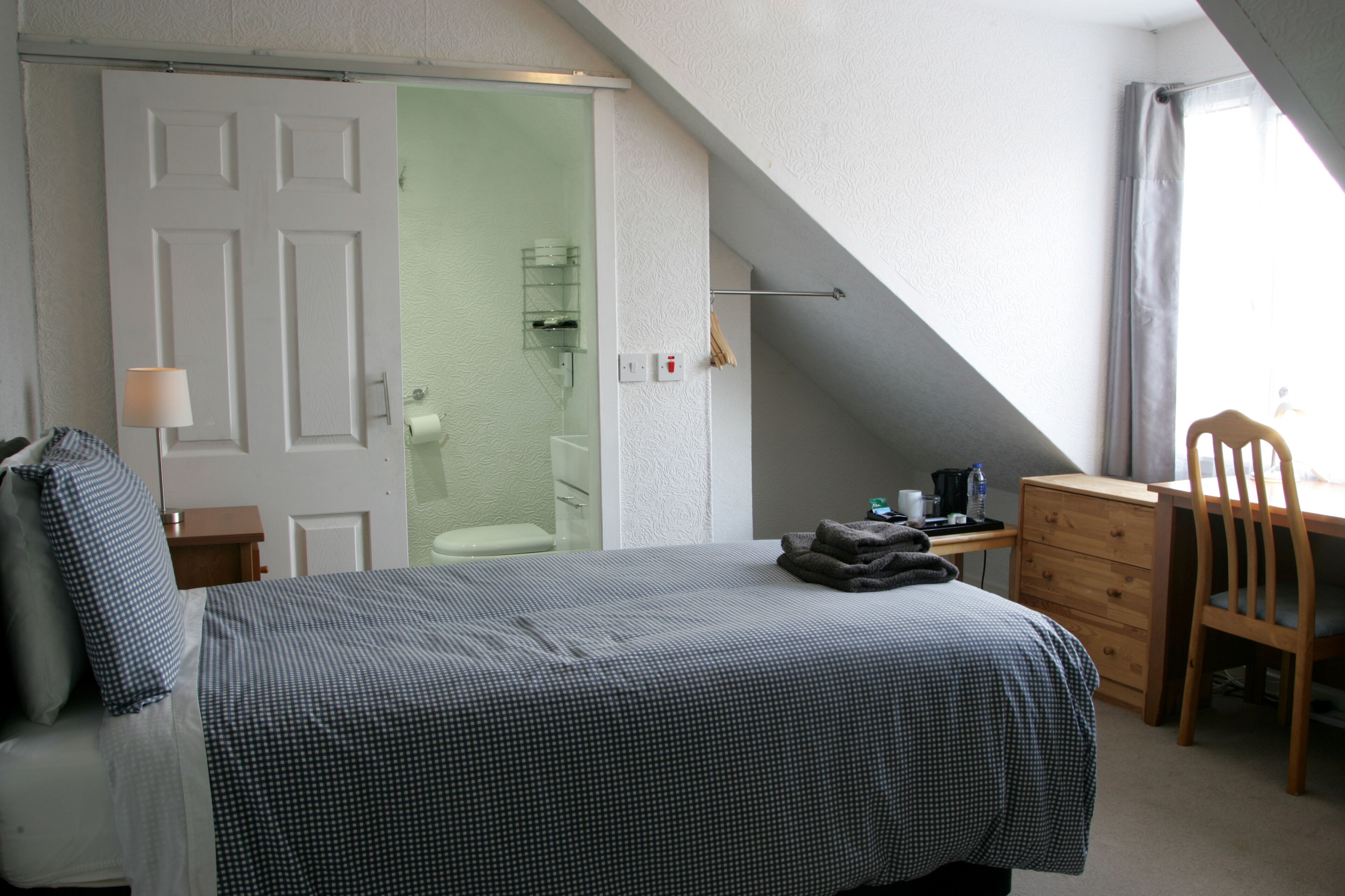 A bedroom with a bed, a desk, a chair, a wardrobe, and a bathroom visible through an open door.