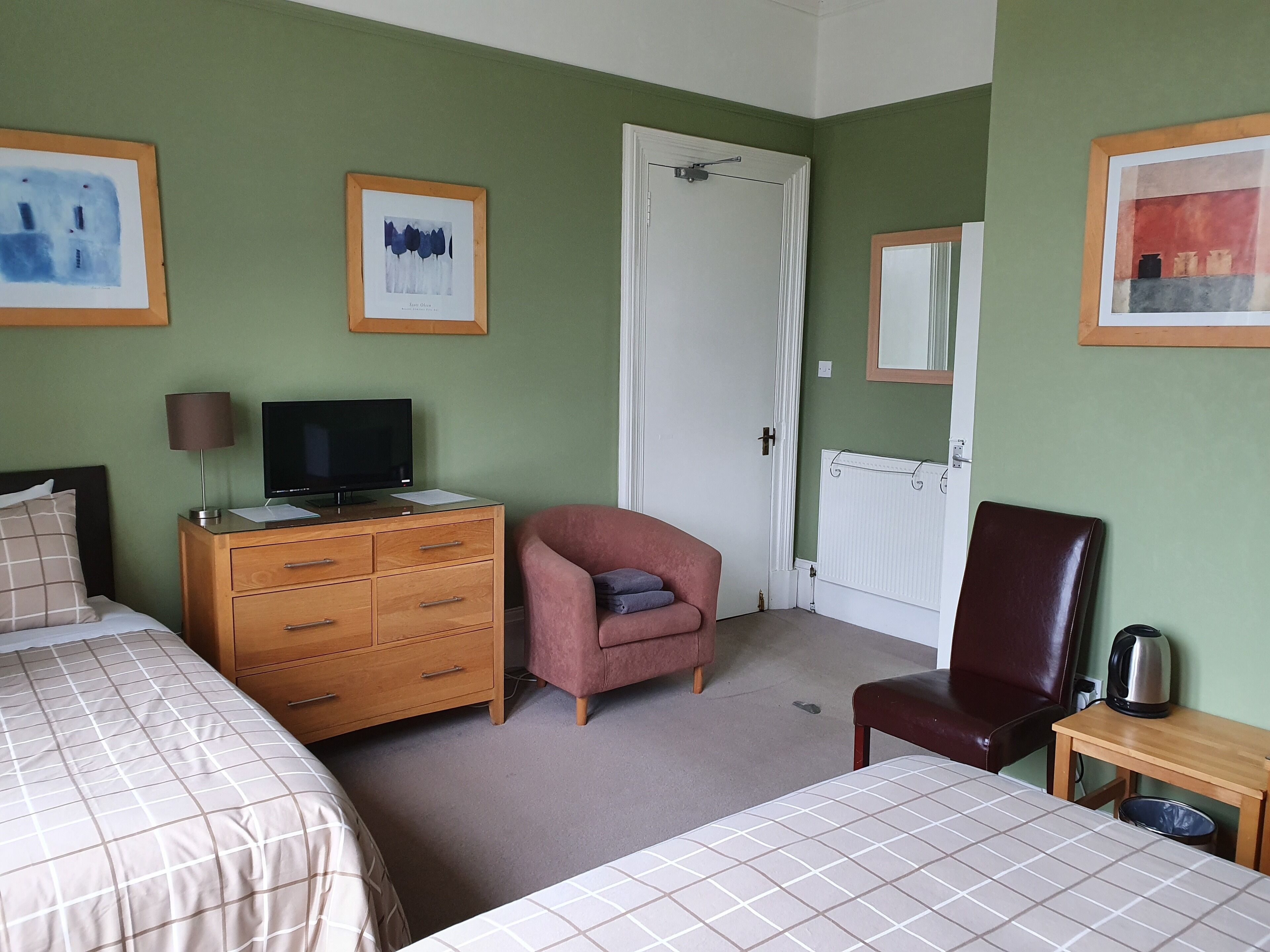 Ein Hotelzimmer mit zwei Betten, einem Holzschrank mit Fernseher, einem pinkfarbenen Sessel, einem braunen Ledersessel und einem kleinen Tisch mit Wasserkocher.
