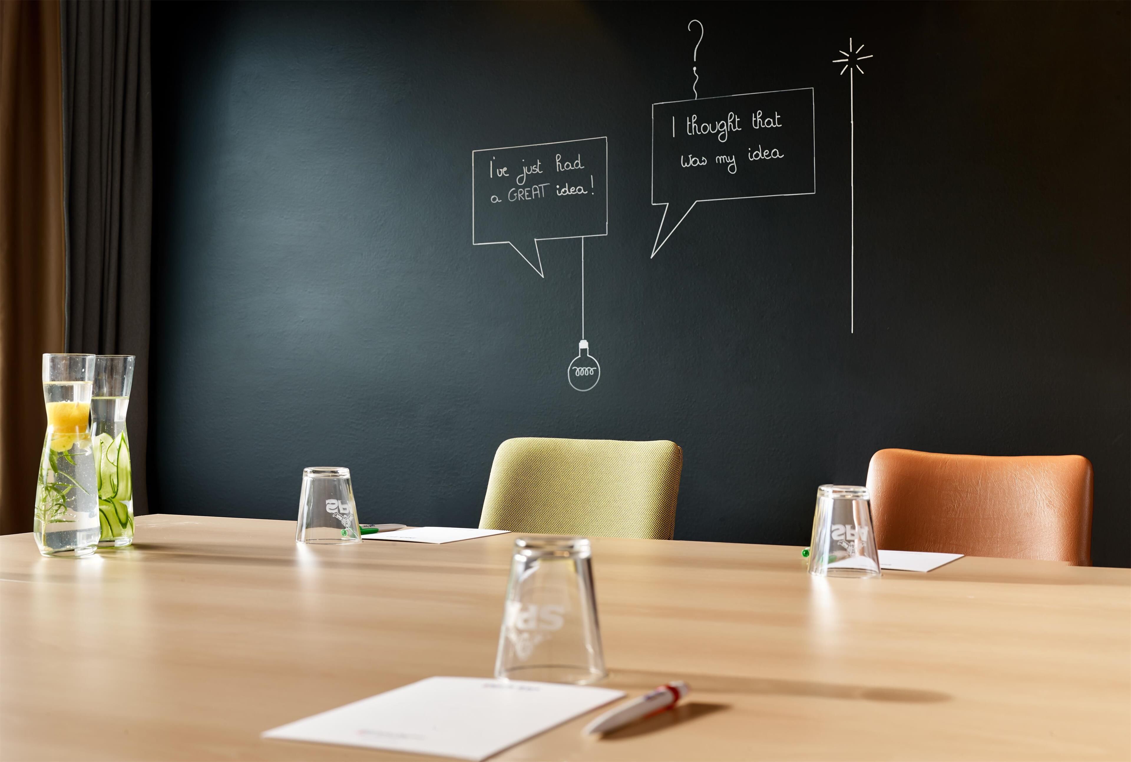 A meeting room with a wooden table, chairs, a blackboard with chalk drawings, and glasses of water.