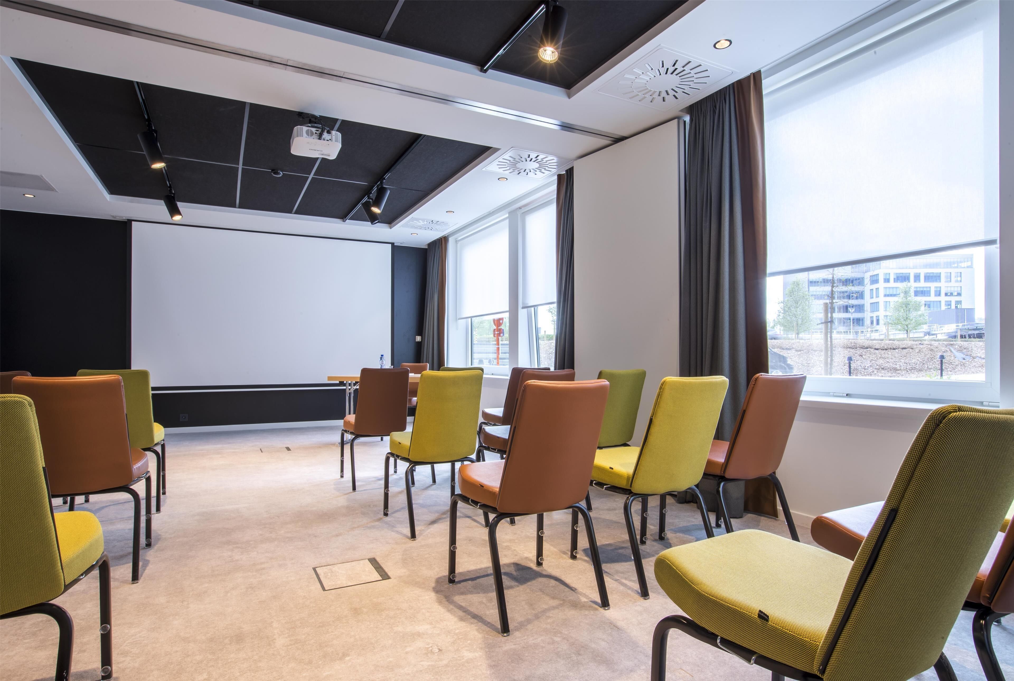 Un salón de conferencias con pantalla de proyector, filas de sillas y ventanas amplias con vistas a edificios.
