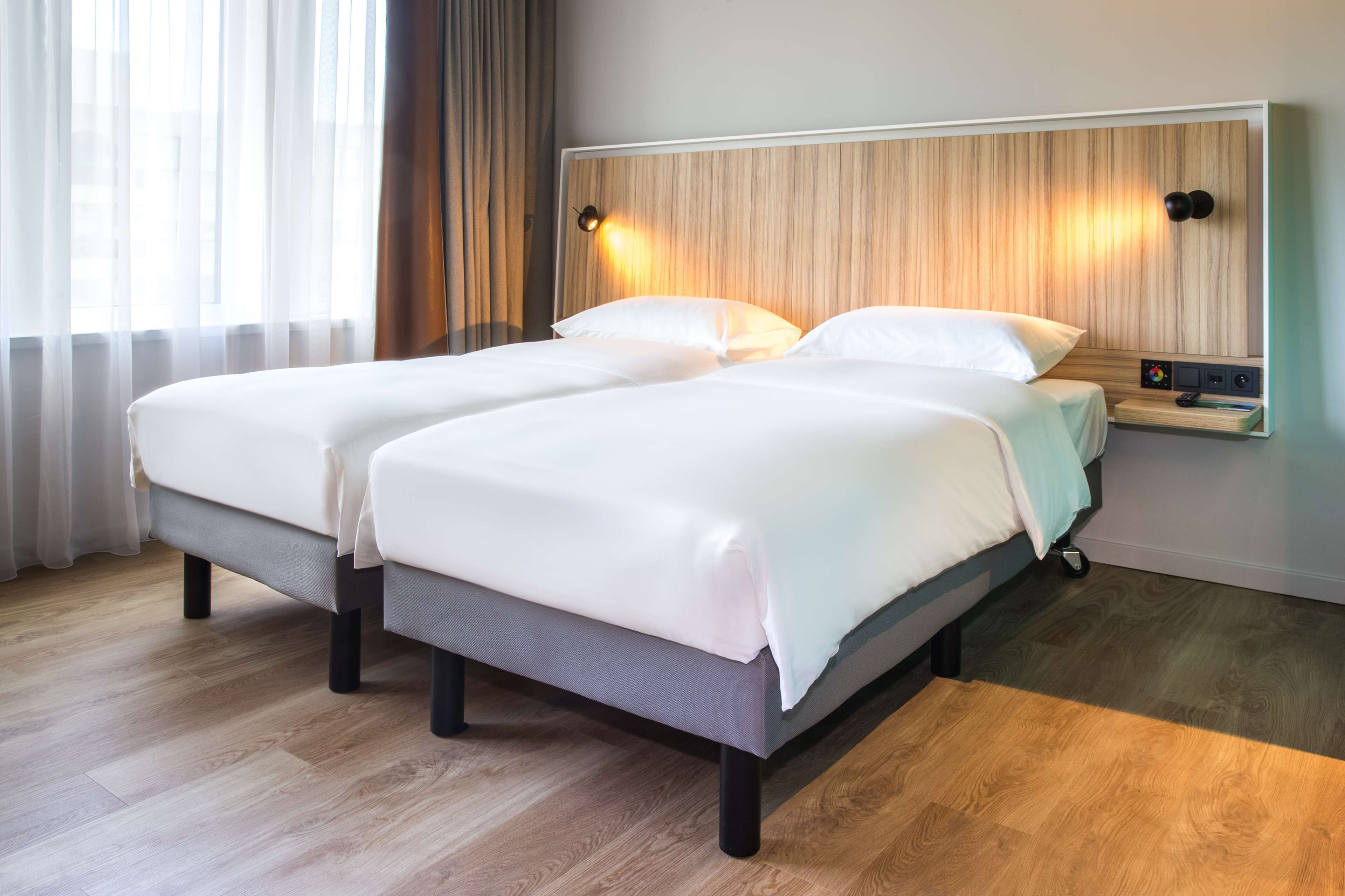 A double bed with a wooden headboard, a bedside table with a lamp, and a view of the city through sheer curtains.