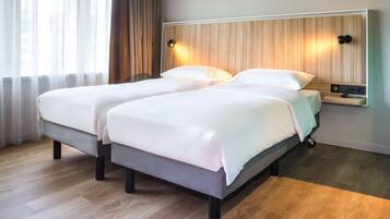 A double bed with a wooden headboard, a bedside table with a lamp, and a view of the city through sheer curtains.