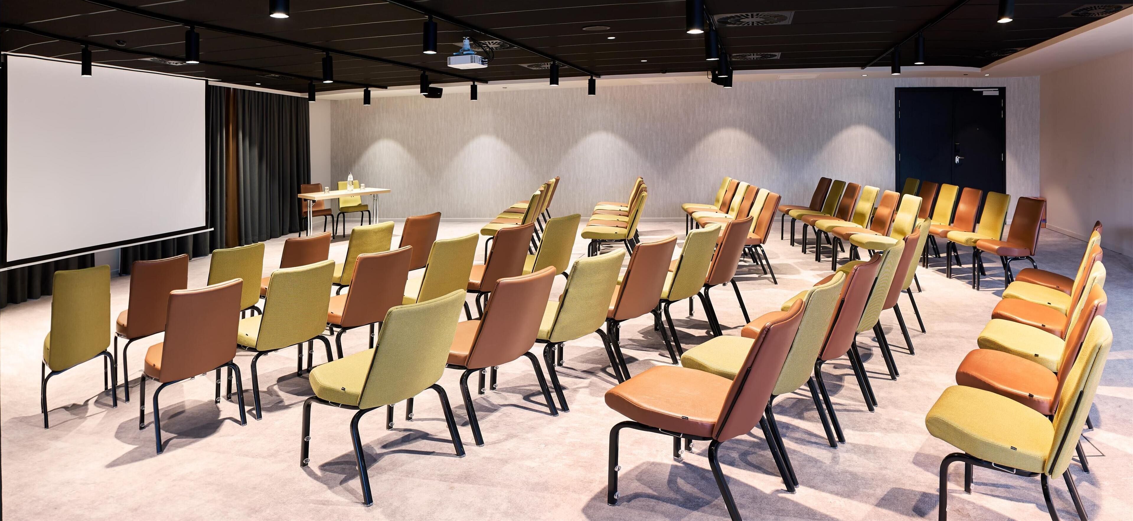 Une salle de conférence avec des rangées de chaises, un écran de projection et une table sur laquelle sont posées des bouteilles d’eau.