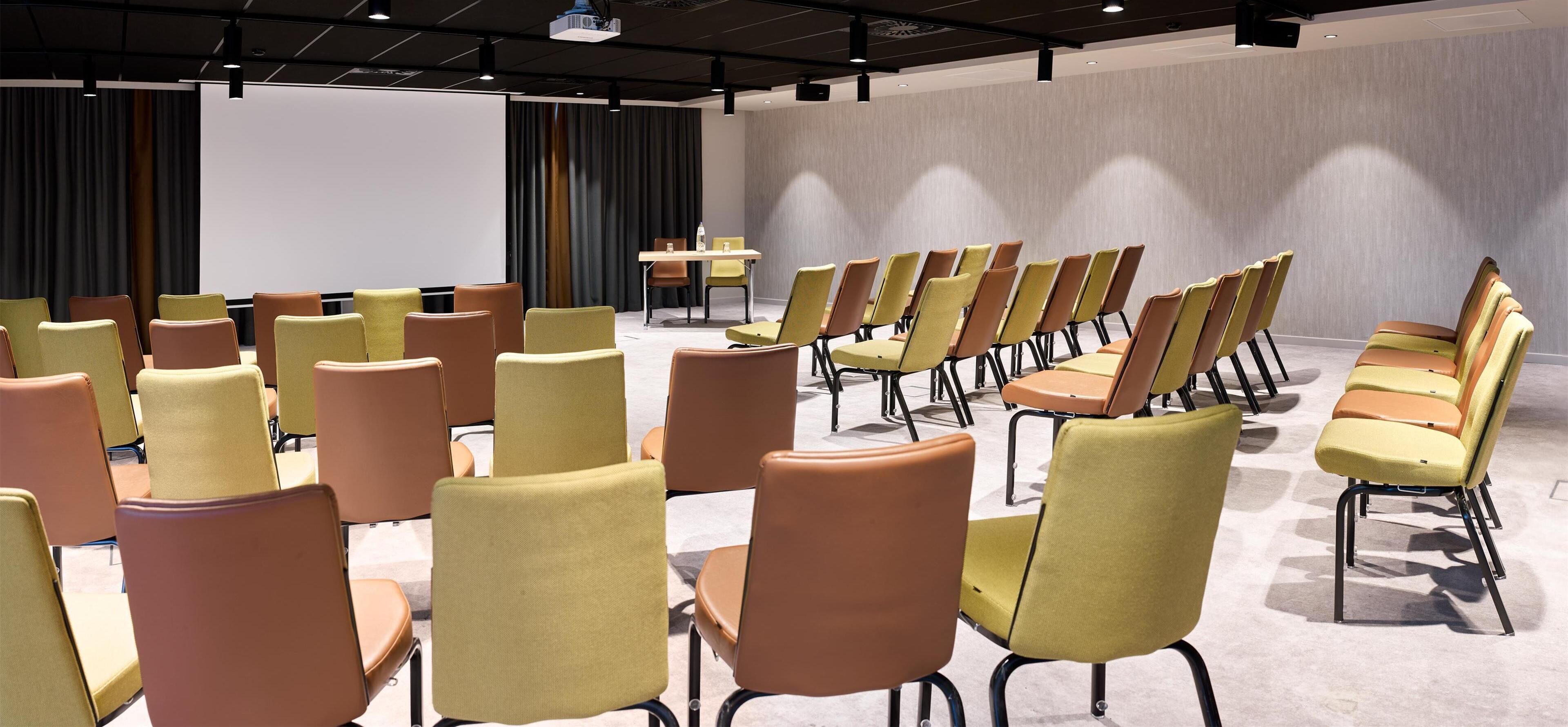A conference room with rows of chairs, a projector screen, and a table with chairs.